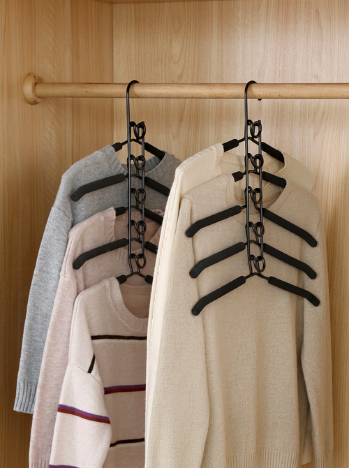 Three black multi-hanger closet organizers hanging on a wooden rod inside a light wood wardrobe, holding gray, pink, and cream knit sweaters.