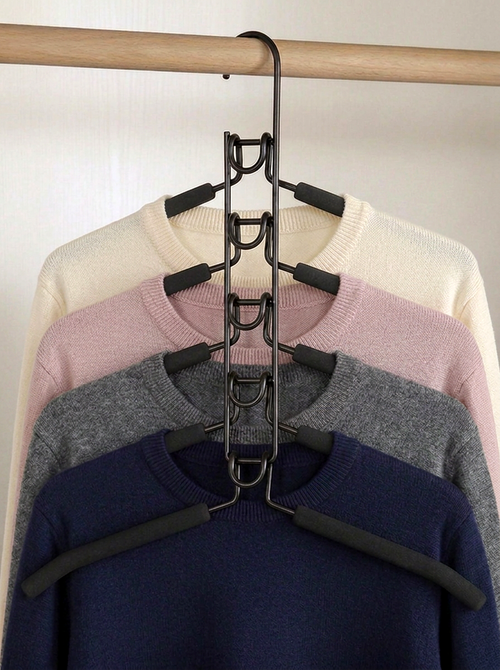 A black metal multi-hanger garment organizer hanging on a wooden rod, holding four nested crewneck sweaters in cream, pink, gray, and navy blue.